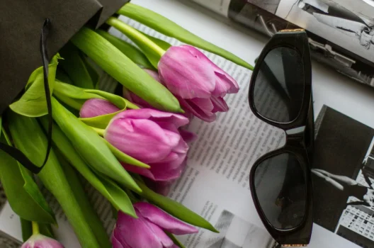 Tendencias de accesorios y complementos de moda para la primavera 2025 Luciendo Gafas de sol negras sobre revistas de moda junto a tulipanes frescos, representando tendencias de accesorios para la primavera 2025.